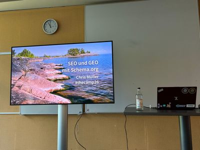 A laptop on a speaker desk next to a presentation monitor. The monitor shows a slide with "SEO GEO mit Schema.org, Chris Müller, #thecämp26"